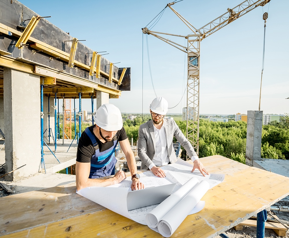 Two people in hard hats working on top of a construction site, with a building being built and crane in the background